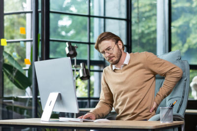 Man at his desk leaning forward in pain holding his back - Spondylolisthesis concept